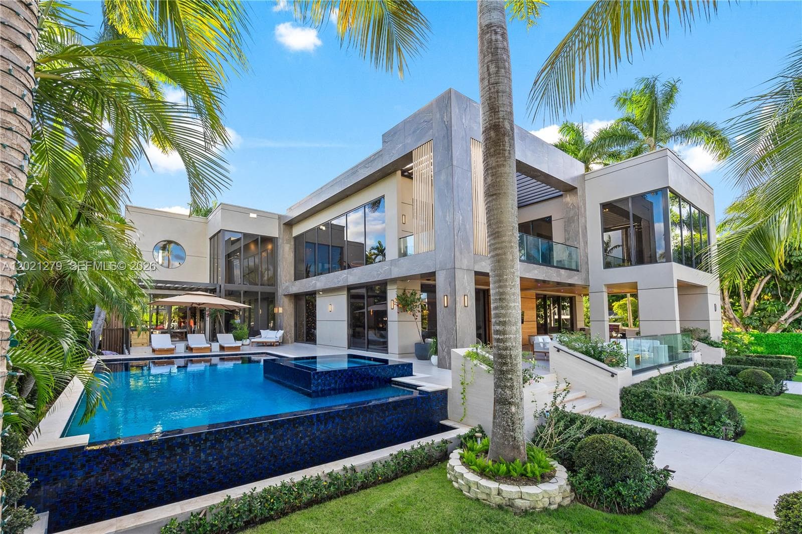 Resort-style pool and spa with dark mosaic tile, sun deck with chaise lounges, pergola, manicured lawn, tropical landscaping, and full two-story modern rear elevation with stone cladding, floor-to-ceiling glass, second-level balcony, and covered loggia.